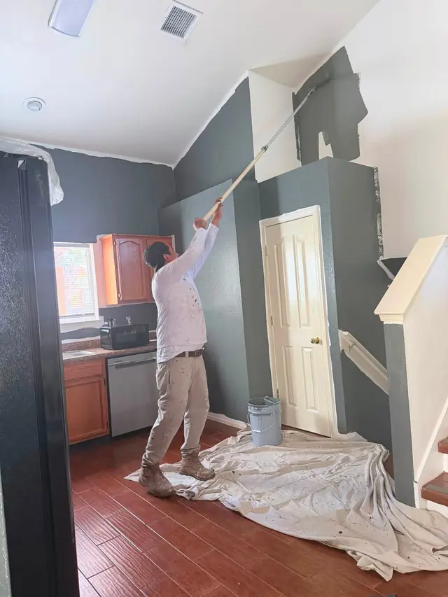 A painter rolling a fresh coat onto a living room wall with the floor covered